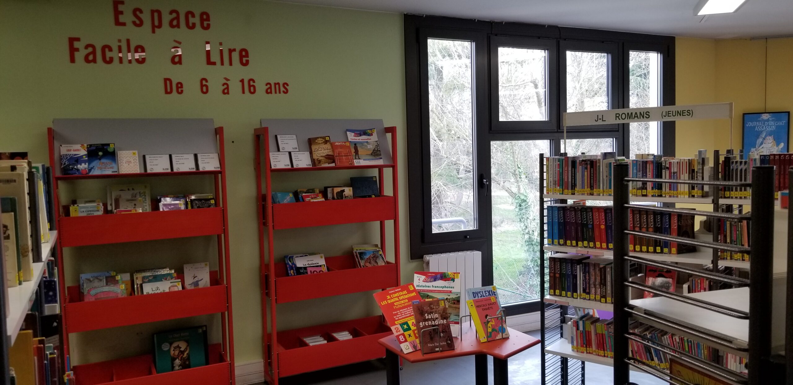 Photo de l'Espace Facile à lire avec mobilier et signalétique rouge qui le rend visible au sein de la bibliothèque. La signalétique indique : Espace Facile à Lire de 6 à 16 ans.